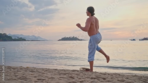 Shirtless martial artist practicing high kicks on sandy beach at sunset, rear view with ocean horizon, calm water, distant boats, islands, warm sky, focused balance coastal fitness