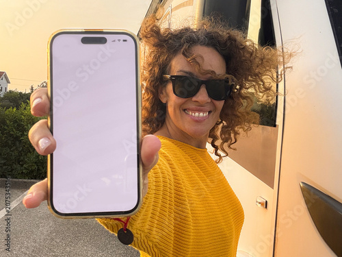 Smiling woman in sunglasses holding a blank smartphone screen toward the camera while standing by a camper van in a sunny outdoor parking area, showing travel, technology and joyful connection
