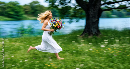 Donna bionda in abito bianco con mazzo di fiori che corre in un prato fiorito, fuoco sul soggetto con sfondo di vegetazione con effetto movimento