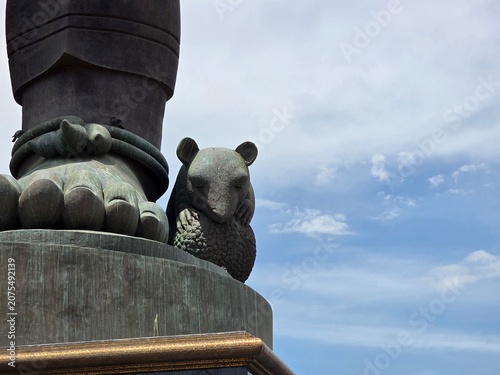 фотографія A tiny bear sculpture nestles against the foot of a colossal statue under a bright, cloudy sky