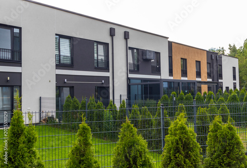 Modern minimalist townhouses with private backyards, dark metal fencing, and decorative green thuja hedge landscaping. Urban