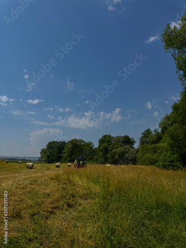 Photos Vertical photo of a tractor producing round hay bales in a wide harvested field on a sunny summer day