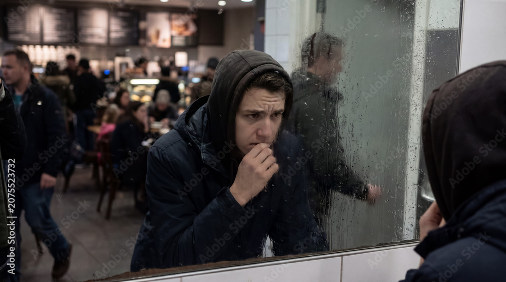 Fototapeta premium Young man wearing a hoodie reflecting in a rainy cafe window, expressing sadness and contemplation while surrounded by city life