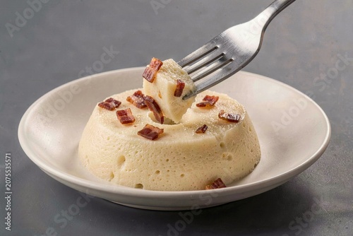 Kugelis in small white bowl, topped with crispy bacon bits. Fork scooping delicious, creamy dessert. Concept of baked potato pudding with bacon onions. 