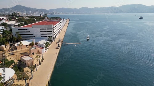 Aerial drone footage moving forward above Acapulco maritime terminal, showcasing the bustling port area, sparkling blue bay water, and distant city landscape under clear skies