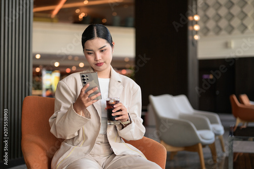 Young Thai Isan Woman Using Phone and Drinking Iced Coffee in Hotel Lobby Copy Space