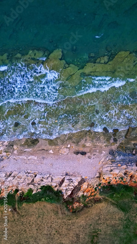 Photography Aerial view of a serene coastline with gentle waves lapping against the rocky shore, transitioning from pebbles to lush greenery