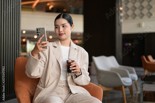 Young Thai Isan Woman Taking Selfie and Drinking Iced Coffee in Hotel Lobby Copy Space