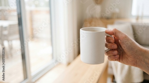 Hand holding a steaming white ceramic mug in a bright minimalist living room, ideal for Website Banner, Hero Header, or Wide Background with Copy Space. Reflects morning wellness and mindfulness.