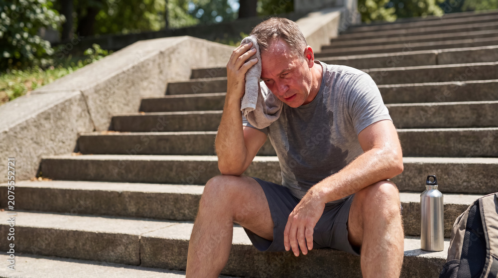 Obraz premium Tired man resting on outdoor stairs after intense workout, wiping sweat, experiencing heat and exhaustion in summer