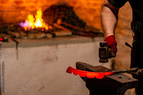 Obraz na plátně Blacksmith hammering a glowing red-hot metal piece on an anvil, forge furnace with bright fire in the background, traditional craftwork inside a dark workshop
