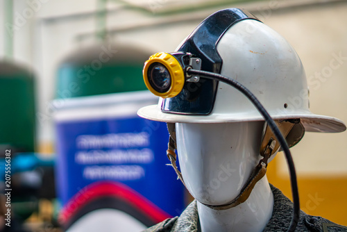 Schilderij op canvas White safety helmet with an attached yellow headlamp worn on a mannequin head, dressed in work uniform, against a blurred industrial interior background with a blue banner and green tanks