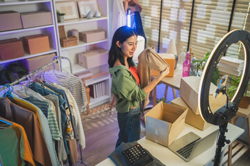 Happy Asian woman looking at camera live streaming fashion clothing sales, online retail e-commerce entrepreneur, social commerce influencer web broadcasting and modern digital side hustle concept