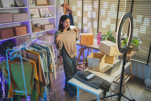 Asian woman entrepreneur presenting clothing product detail, online retail e-commerce merchant live streaming sales, small business owner working at home office and social commerce concept
