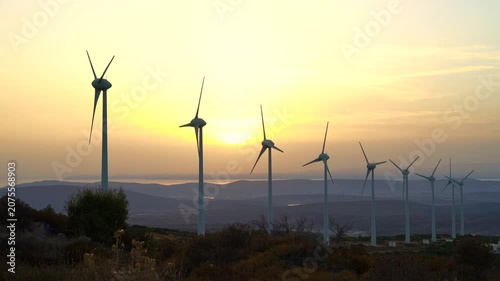 A row of wind turbines stands on a hillside as the sun sets behind them, generating clean energy. The turbines' white blades rotate in the breeze, surrounded by sparse vegetation.