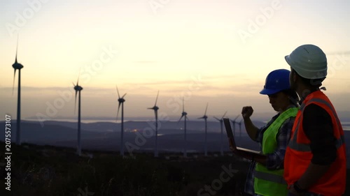 Two workers in safety gear, one wearing a blue hard hat and high-visibility vest, the other in a white hard hat and orange vest, examine wind turbines on a hillside at dusk.