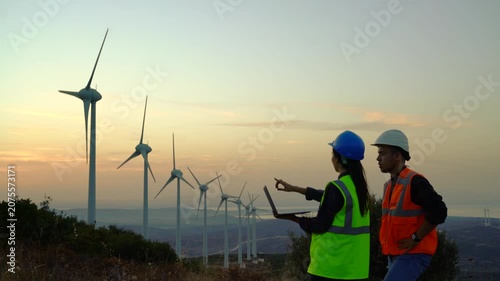 Two workers in safety gear, one wearing a blue hard hat and high-visibility vest, the other in a white hard hat and orange vest, examine wind turbines on a hillside at dusk.
