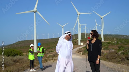 Man in traditional Middle Eastern attire interviews woman with microphone at wind farm
