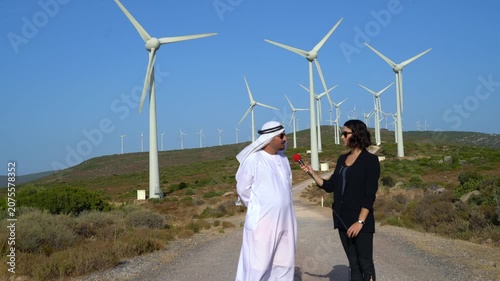 Man in traditional Middle Eastern attire interviews woman with microphone at wind farm