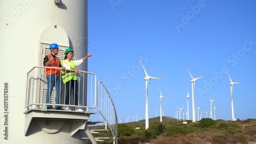 The two engineers, are standing on a platform attached to a wind turbine tower, inspecting the surrounding wind farm landscape with several turbines visible in the background.