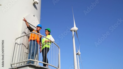 The two engineers, are standing on a platform attached to a wind turbine tower, inspecting the surrounding wind farm landscape with several turbines visible in the background.
