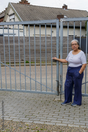 Senior woman grasps handle of closed metal gate blocking access to rural street, trying to open it, house and a wooden in background, long, gray hair tied back and is dressed casually