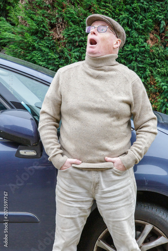 Older man with an expression of astonishment or surprise on face looks upwards, leaning against body of blue car, hands in pockets, green trees in background, wearing casual clothes