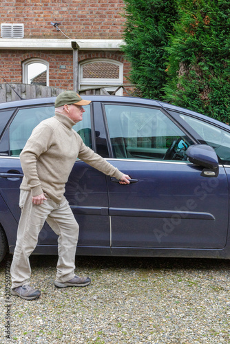Older man in action to open the car by grabbing handle, car parked on  rural street with brick house and green trees in background, casual clothes, cap, pants and brown turtleneck sweater