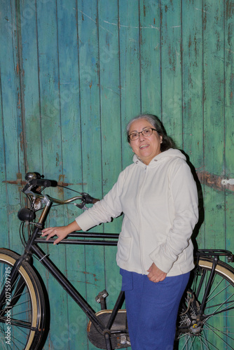 Smiling older woman standing next to a bicycle, looking at camera, wearing casual clothes, white sweater, blue pants, no makeup and gray hair, worn green wooden wall in background