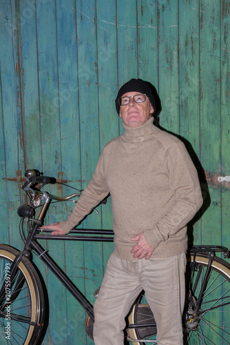 Older man standing next to bicycle looking intently upwards, casual clothes, black winter hat, turtleneck sweater and brown pants, worn green wooden wall in the background