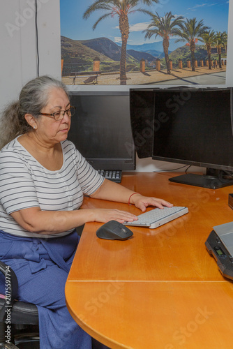 Elderly woman typing on computer keyboard with serious expression, seated in chair in home office, wearing casual clothes, long gray hair tied back. Concept of senior citizens working from home