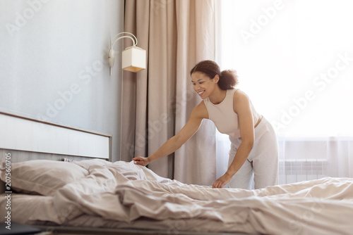 Foto Happy African American woman making bed in bright bedroom, smoothing sheets and smiling