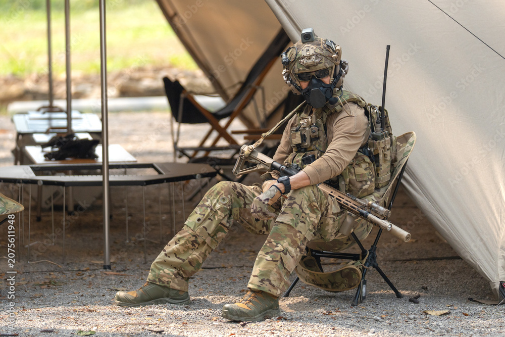 Fototapeta premium One army man or soldier sit on chair and hold his weapon in front of military tent with look at camera.