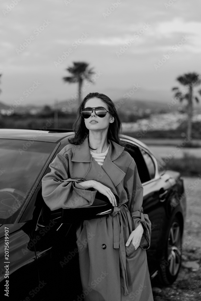 Naklejka premium A stylish woman stands beside a sleek car in a trench coat and sunglasses, exuding confidence. A street lifestyle moment with fashion, independence, and bold attitude.