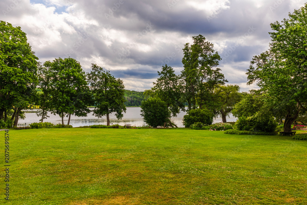 Fototapeta premium Beautiful view of lakeside lawn bordered by trees beneath dramatic clouds over calm water. Sweden.