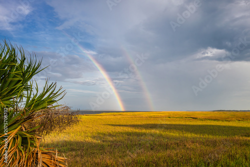 Two rainbows appear over a field of grass under a cloudy sky during the late afternoon hours near the coastline