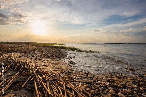 Sunset by the river shore with grass and stones in the foreground during evening hours