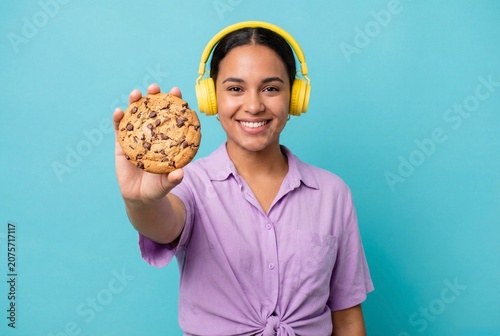 Young smiling woman in yellow headphones holding huge chocolate chip cookie against blue background