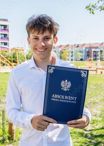 Happy Polish boy in white shirt holding elementary school graduation certificate book outdoors
