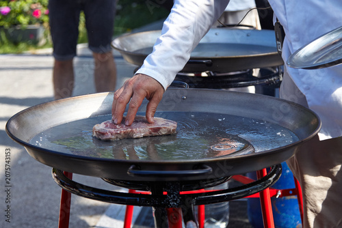 Schilderij op canvas Chef placing raw steak onto large steel grill with water, surrounded by outdoor