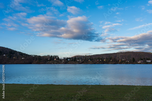 A serene view of Zivohost village across the Vltava river with hills in the background. The bright blue sky with soft clouds enhances the peaceful atmosphere, creating a sense of calm.