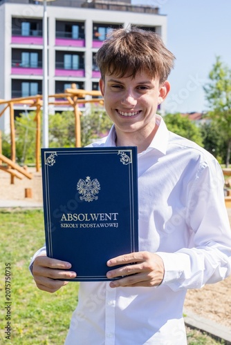 Happy Polish boy in white shirt holding elementary school graduation certificate book outdoors