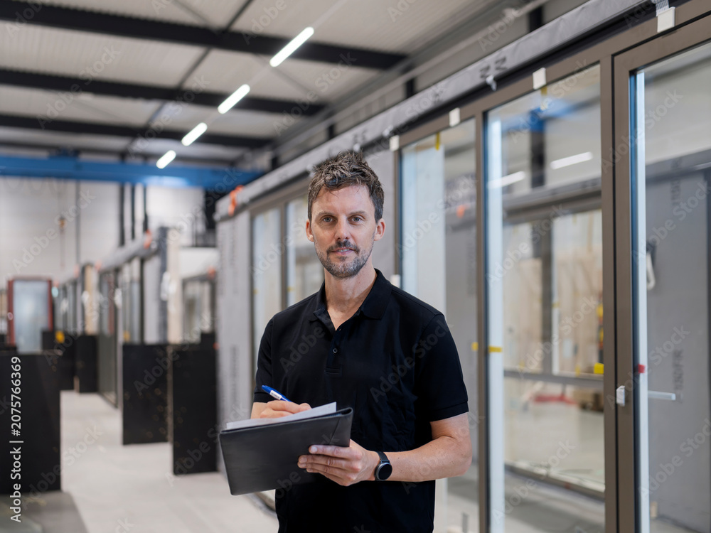 Fototapeta premium Quality worker stands centred writing on a clipboard among glazed door units, a neutral focused look in a production hall.
