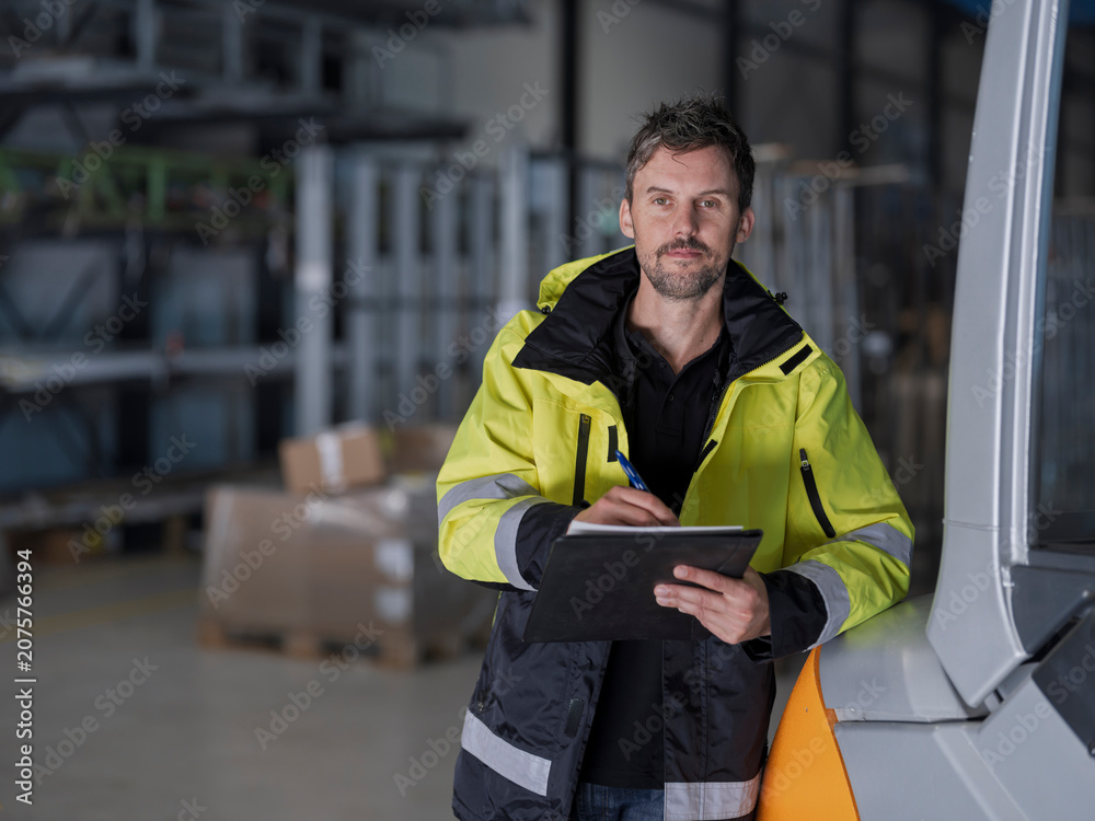 Fototapeta premium Hi-vis warehouse worker records details on a clipboard beside a forklift, calm and neutral with open floor to the side.