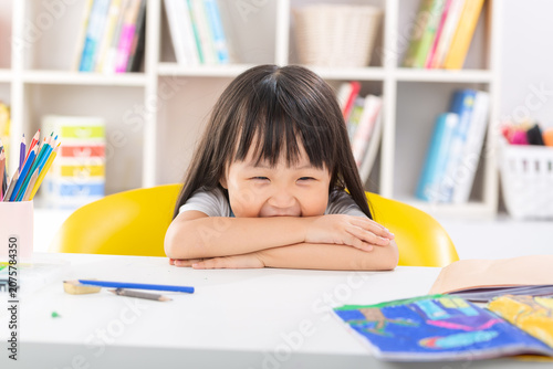 Obraz na plátně A shy and playful young girl burying her face in her arms on a desk while giggling happily in a bright classroom