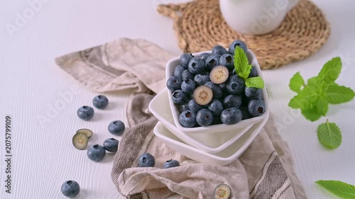 A white square bowl filled with plump blueberries sits on a rustic linen cloth, featuring one berry sliced in half to show its creamy interior under bright natural light.