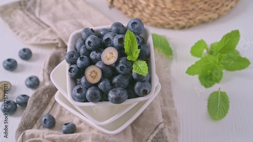 A square bowl filled with ripe, plump blueberries features one berry cut in half to show its creamy interior. The rustic composition includes a textured linen cloth against a bright, airy background.