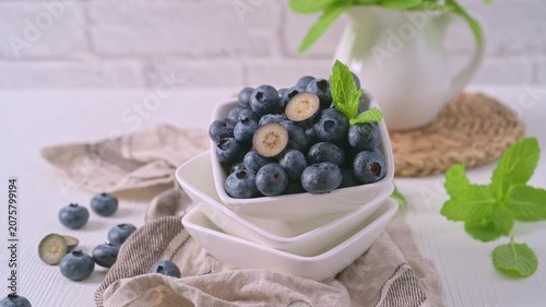 A white square bowl filled with plump blueberries sits on a rustic linen cloth, featuring one berry sliced in half to show its creamy interior under soft, bright lighting.