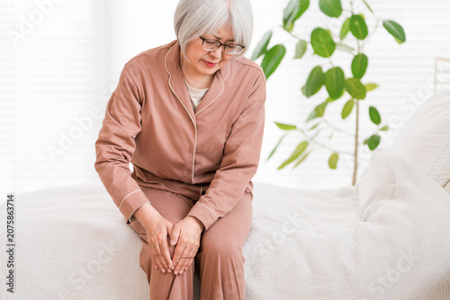 Foto A senior woman in pajamas sits on her bed after waking up, complaining of knee pain (knee, knee pain)