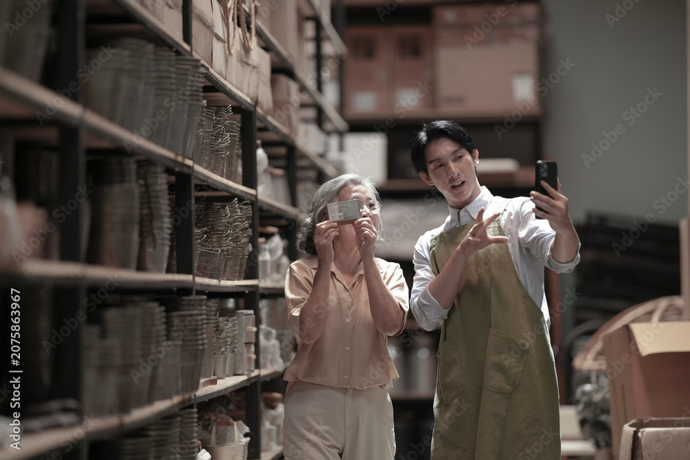 Fototapeta premium Asian shop owners in aprons inspecting handmade products stored on tall shelves among rows of inventory. Showing pride in locally made products. Independent retail and craftsmanship concept.